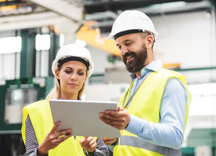 A male with a beard and young female sharing a computer wearing high-vis and hard hats in a manufacturing setting.